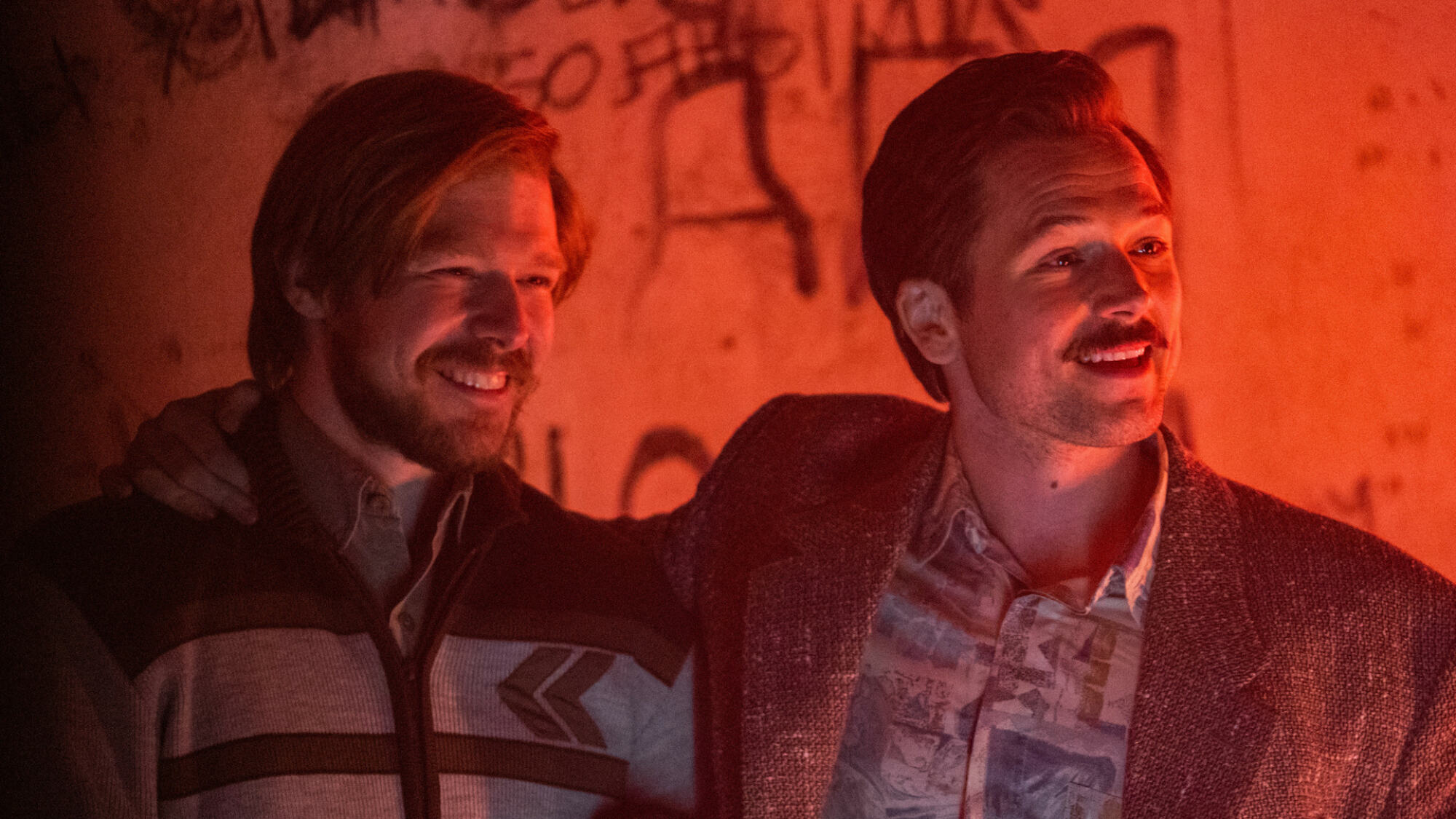 Two men with beards and mustaches stand together ina. red-lit room in "Tetris"