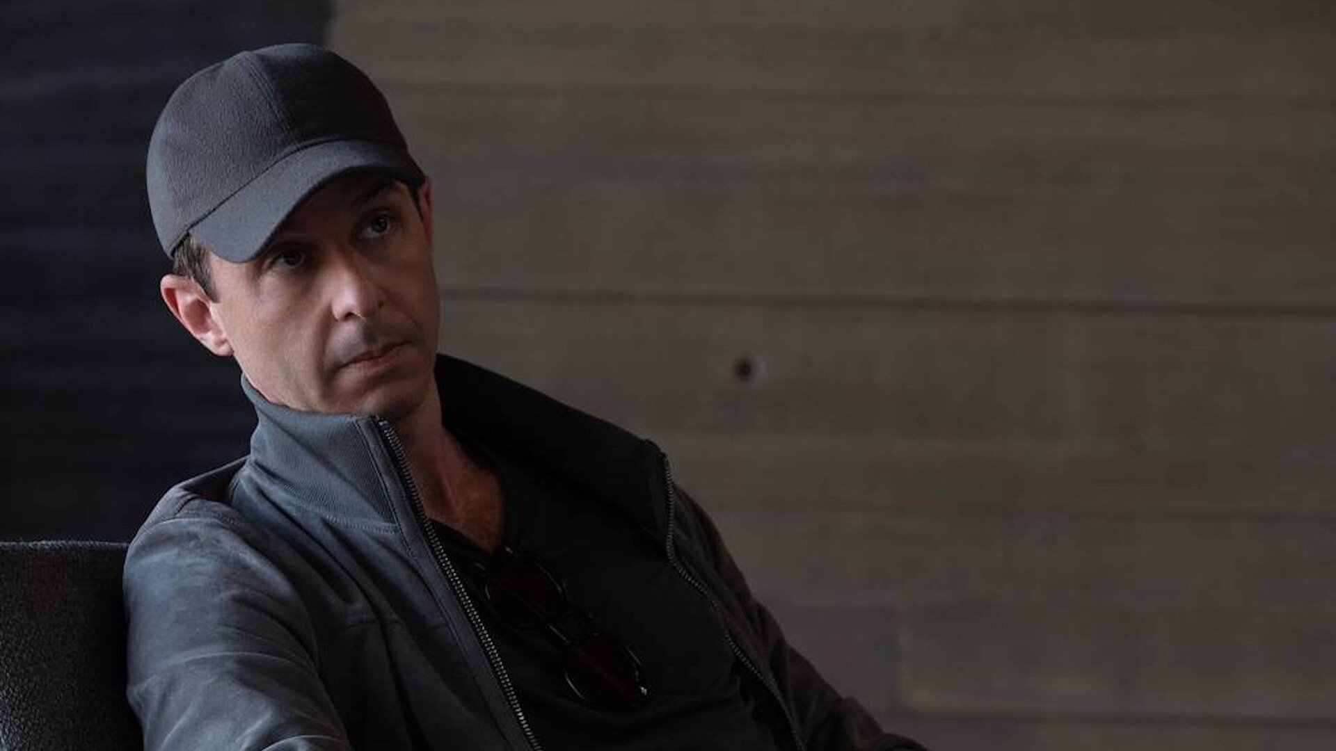 A man wearing a black cap and jacket sits on a chair. 