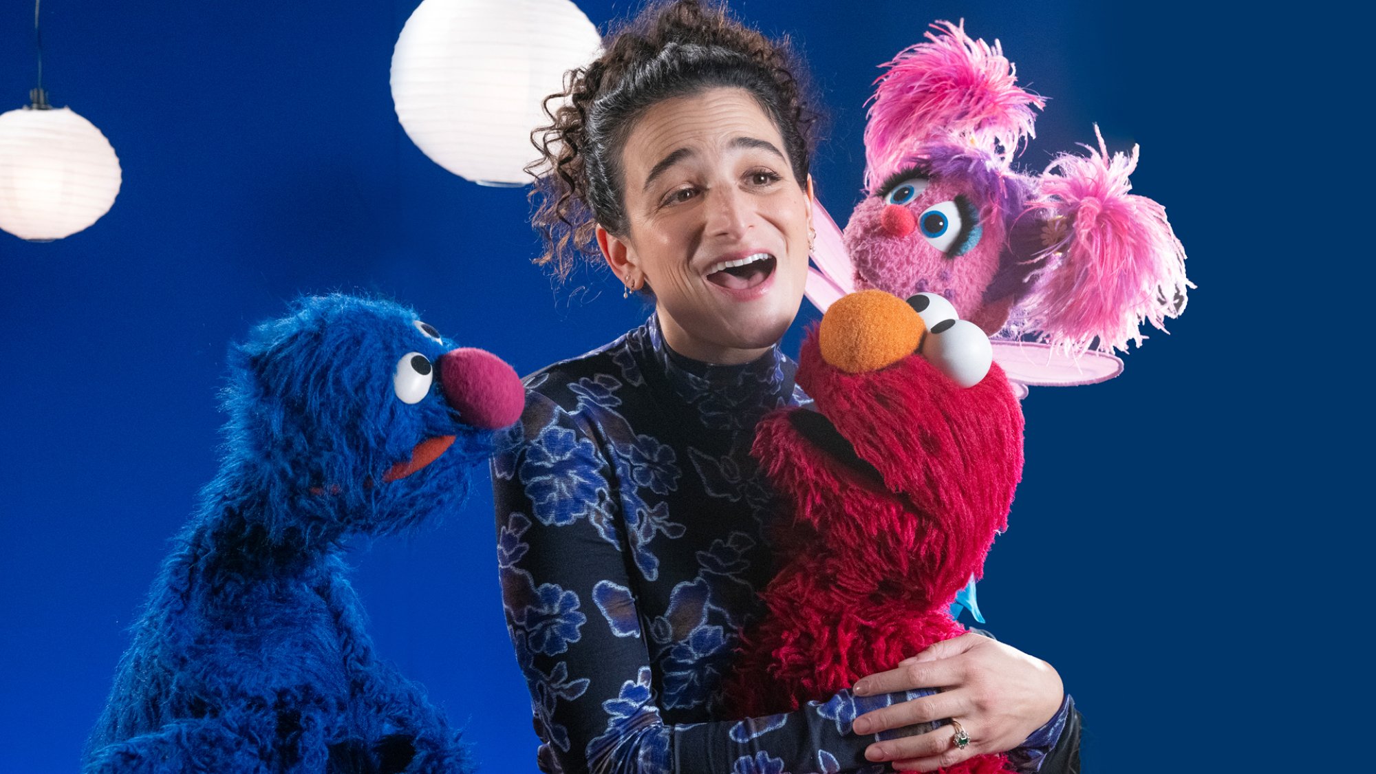 Jenny Slate holds onto Elmo and Abby while Grover watches. They are standing in front of a blue wall.