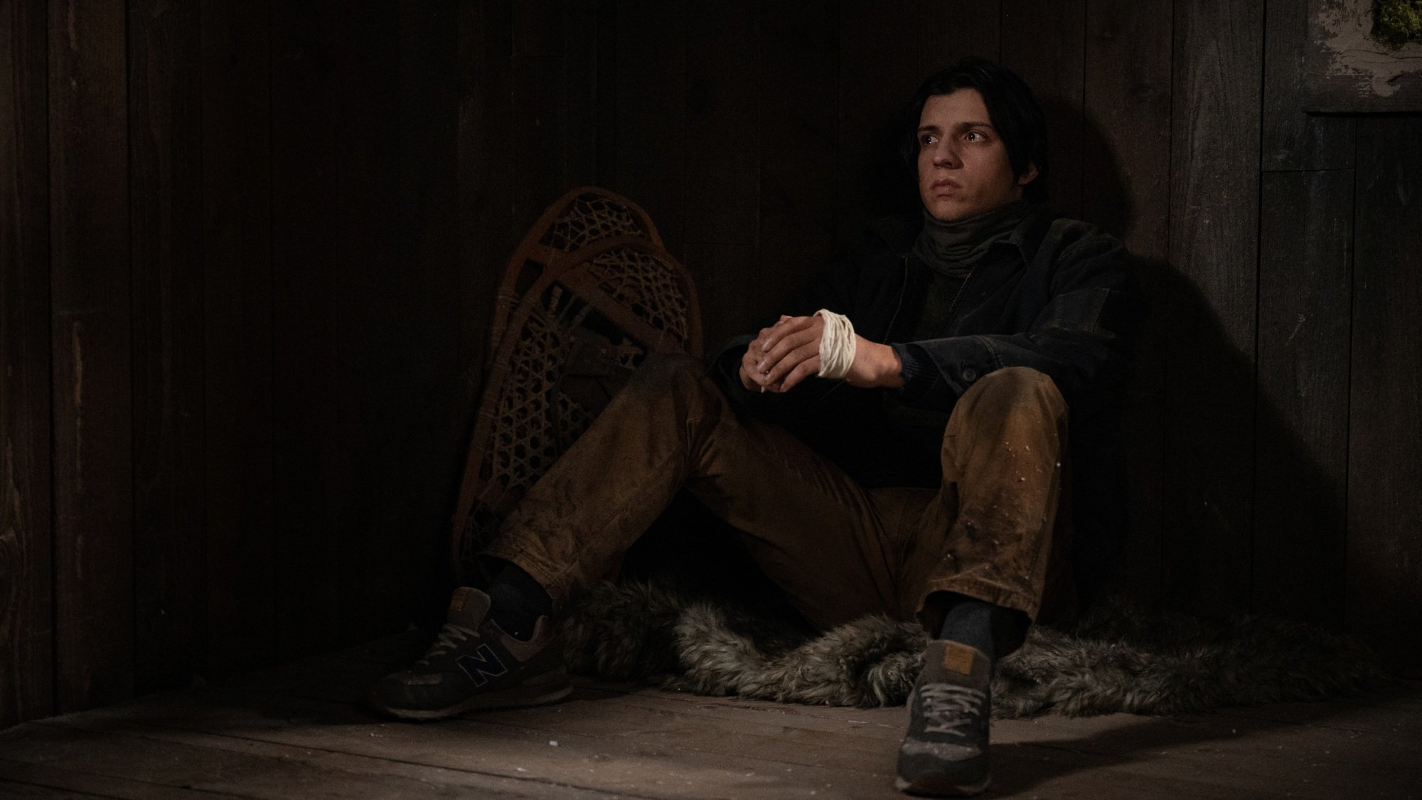 A young man in winter clothes sits in the corner of a cabin.