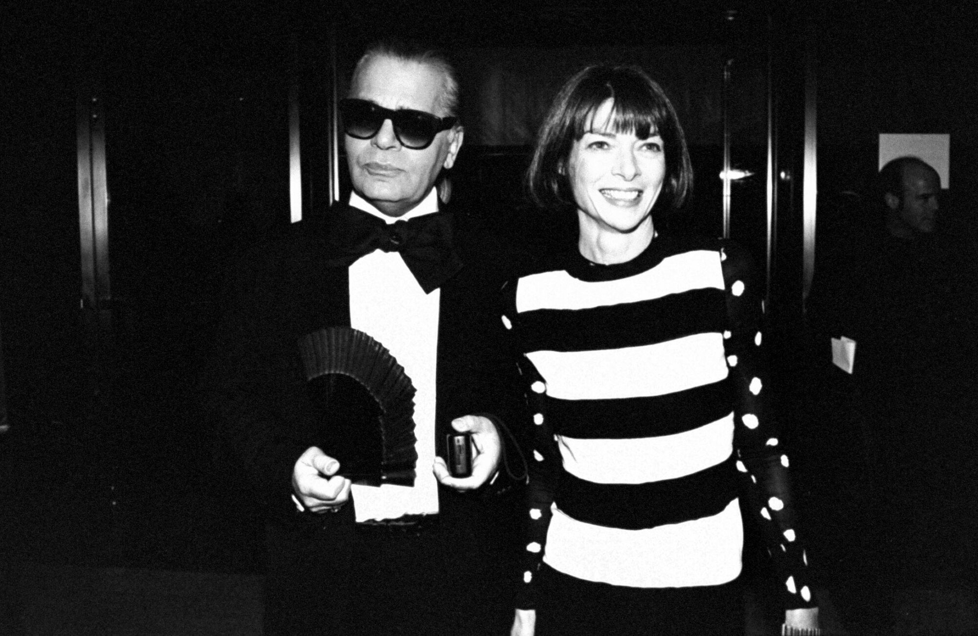 Black and white photo of Lagerfeld, wearing a tuxedo with a large bow tie and holding a hand fan, arm-in-arm with Wintour who is wearing a shirt with large horizontal stripes.