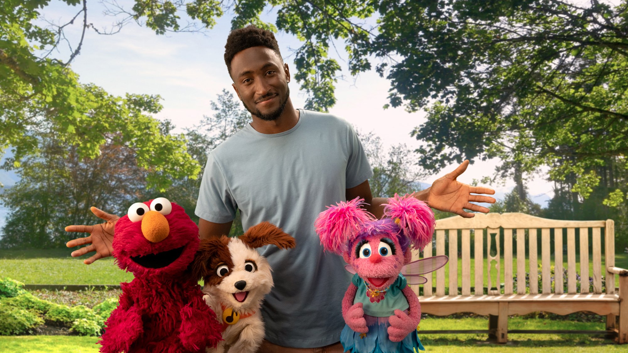 Marques Brownlee, Elmo, Abby, and Tango the dog stand in front of a bench in a sunny park.