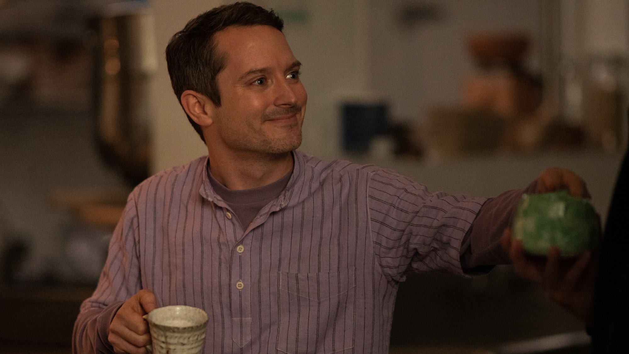A man in a purple button-up shirt offers someone a green mug.