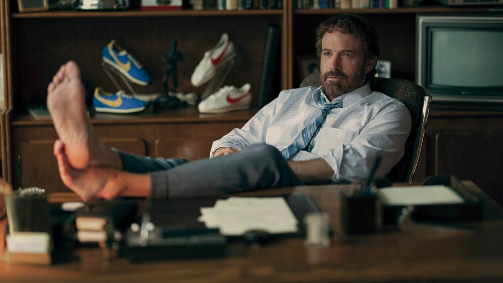 A man in a shirt and tie reclines in his office with his bare feet on the desk in the movie "Air."