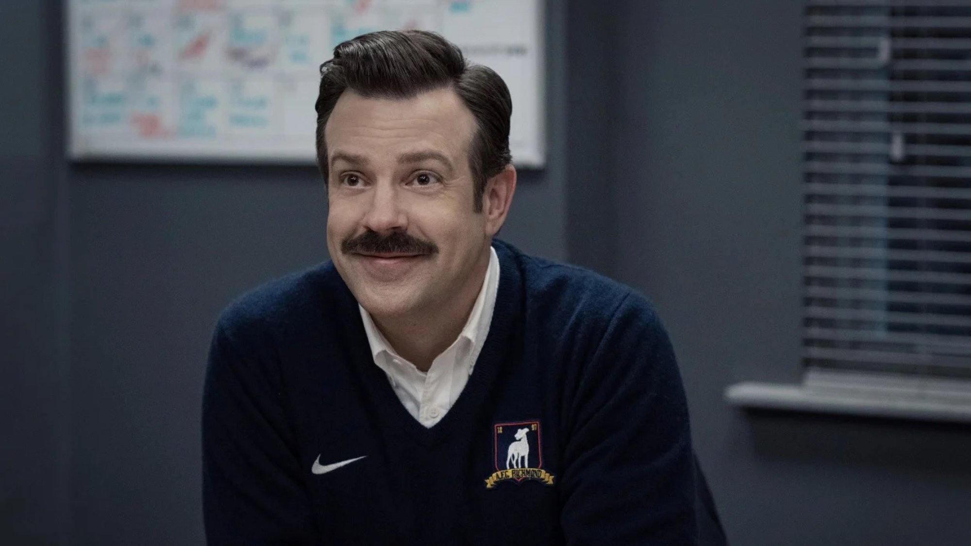 A man with a moustache sits in a football locker room smiling on the TV show "Ted Lasso"