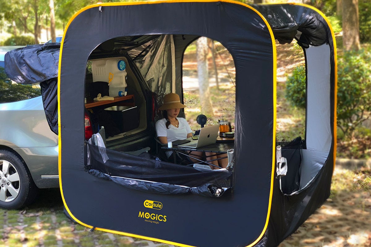 A woman sitting inside a CARSULE Pop-Up Cabin attached to her car and working on a laptop.