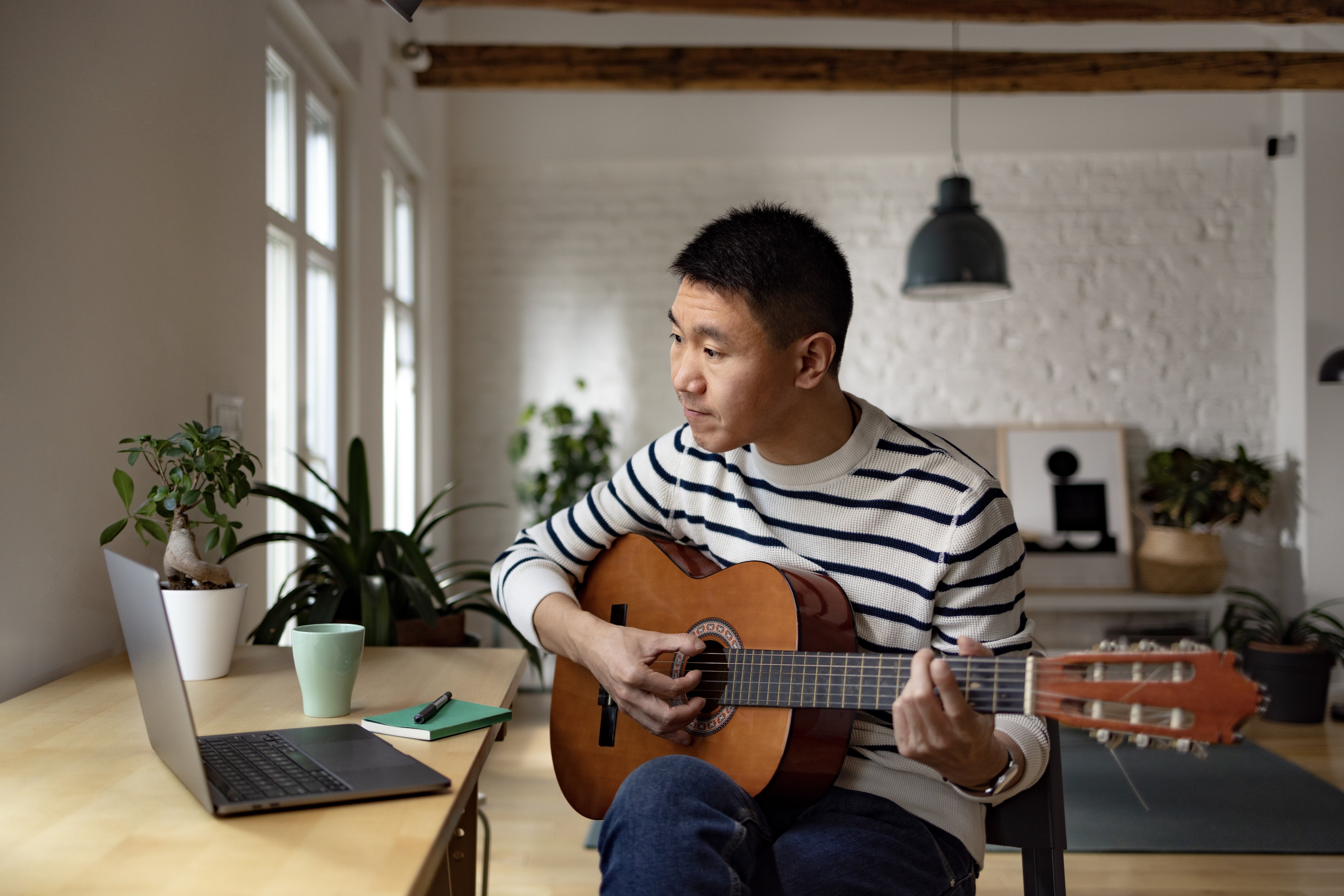 Man taking a virtual guitar lesson, playing an acoustic guitar while sitting in front of an open laptop.