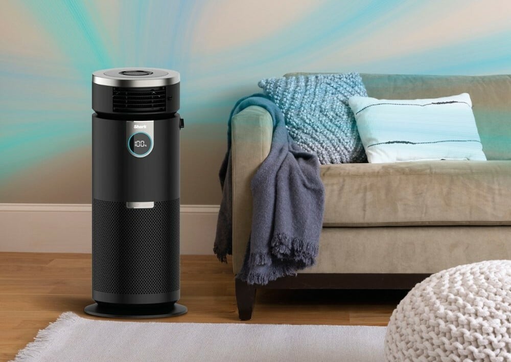 a shark air purifier sits to the left hand side of a beige sofa that has a blue throw blanket over the left arm.