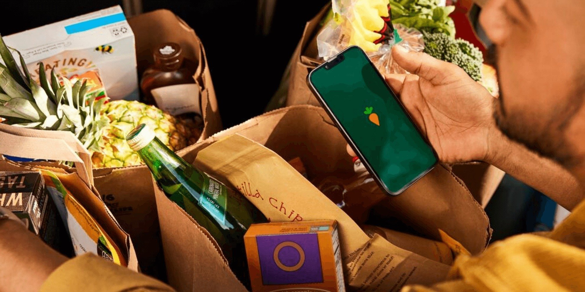 a person looks at their phone with the instacart carrot on the screen while standing over many bags full of groceries