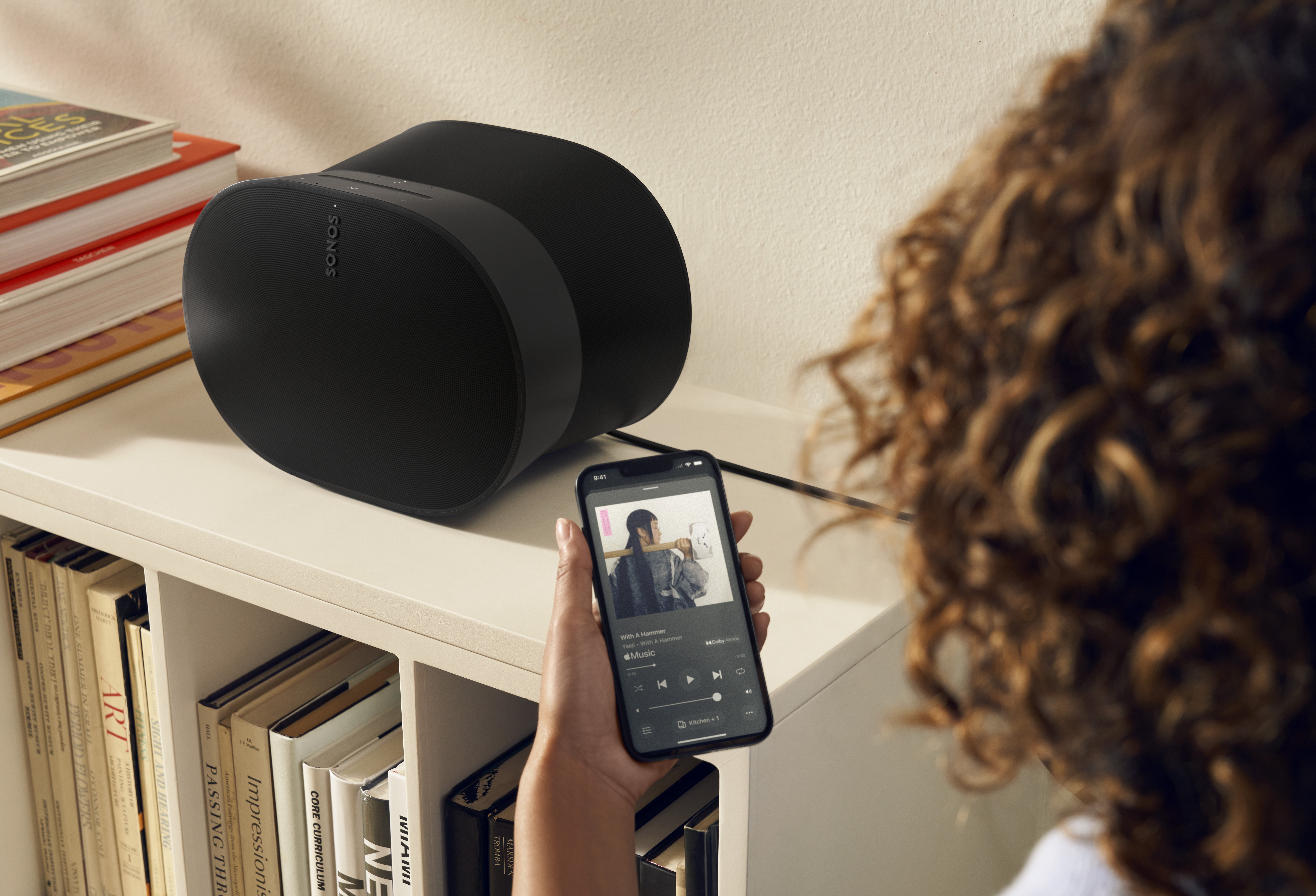 A person points their Sonos app shown on a smartphone at a Sonos speaker sitting on a shelf.