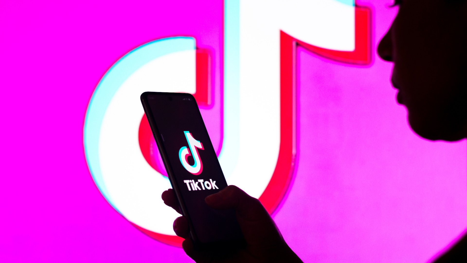  In this photo illustration, a woman's silhouette holds a smartphone with the TikTok logo displayed on the screen and in the background.