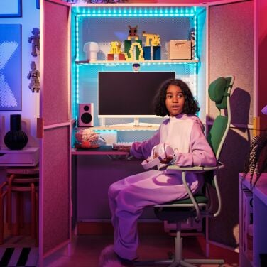 A girl sits in a green gaming chair in front of a large cabinet whose doors are open and reveal a PC gaming set up inside.
