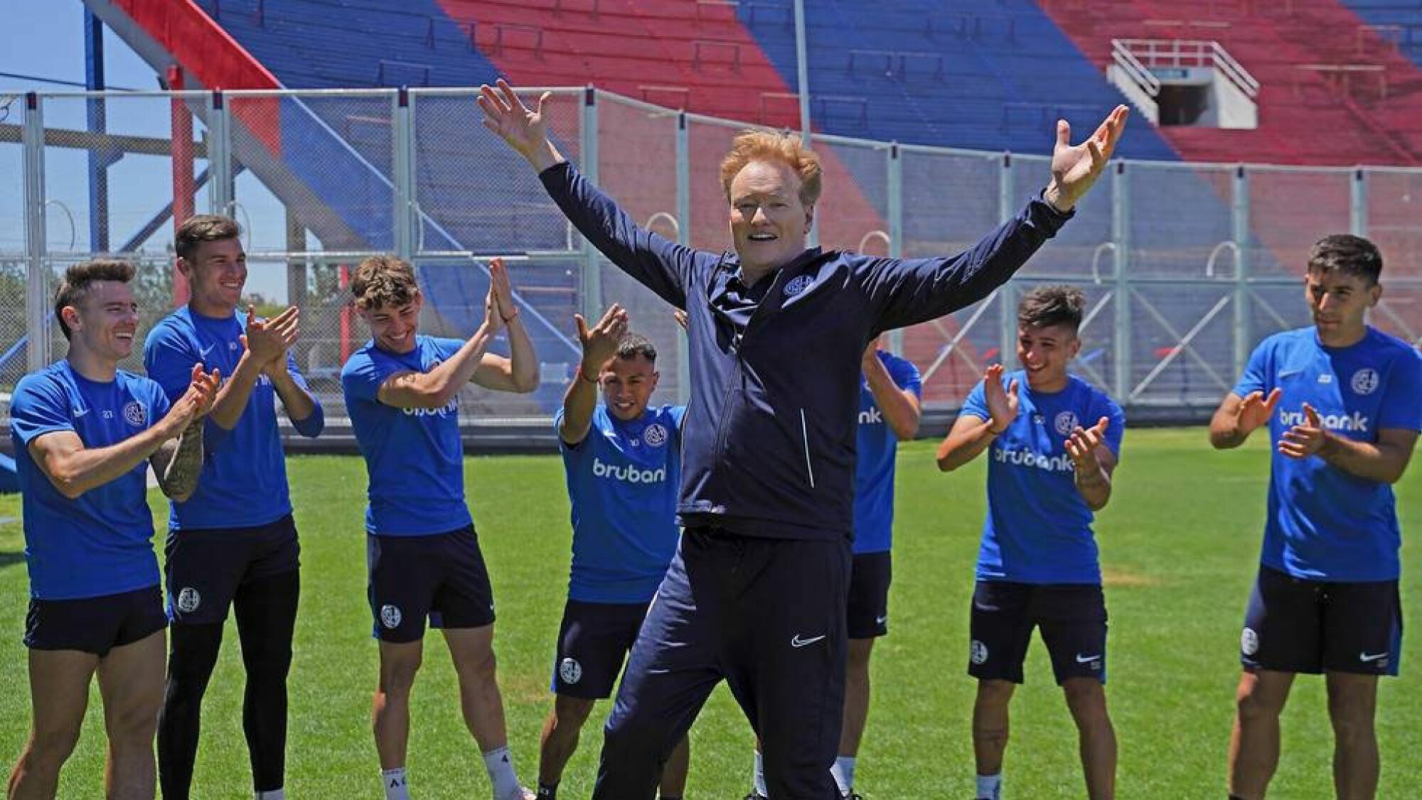 A man stands on a football pitch celebrating in front of other men who clap.