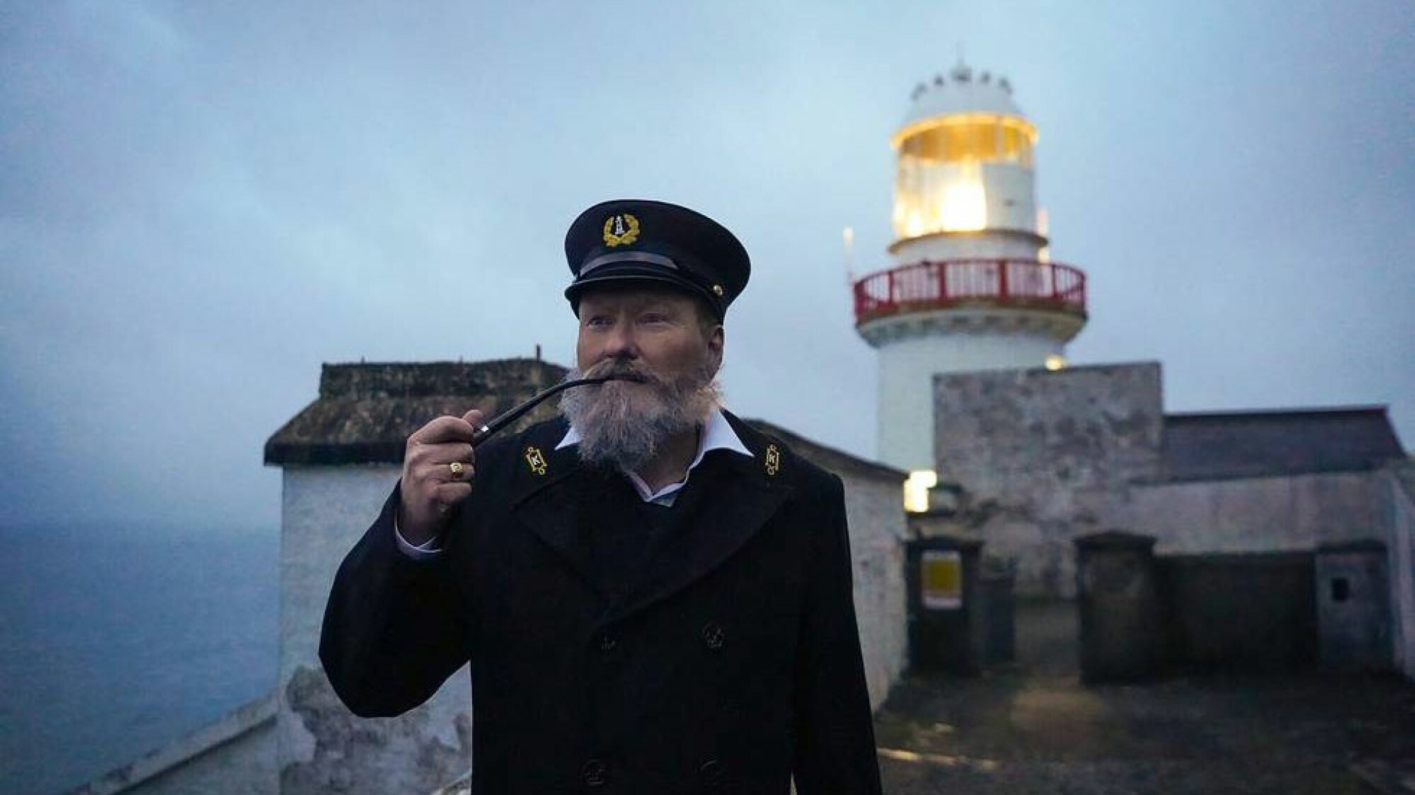 A man in an old lighthouse keeper outfit stands in front of a lighthouse.