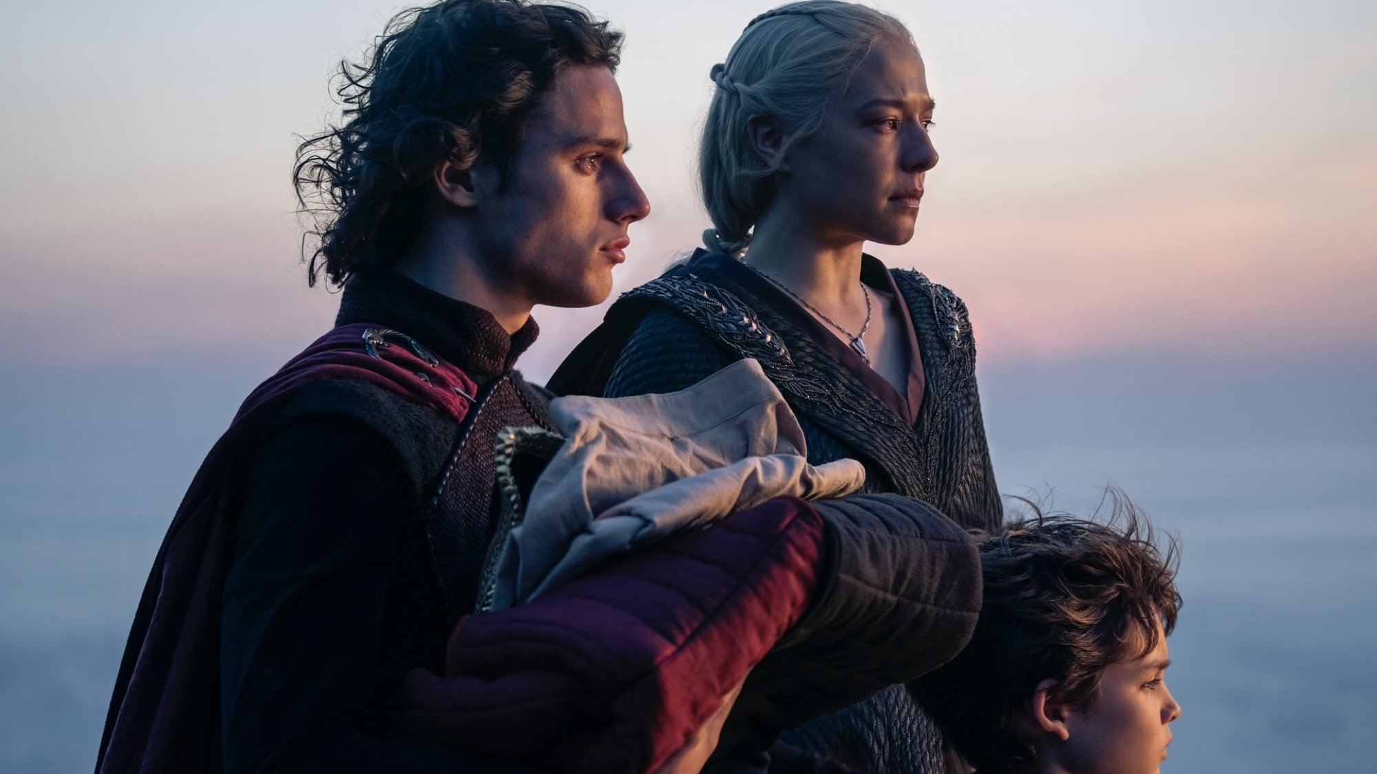 Jacaerys, Rhaenyra, and Joffrey stand together with their backs to the sea at sunset.