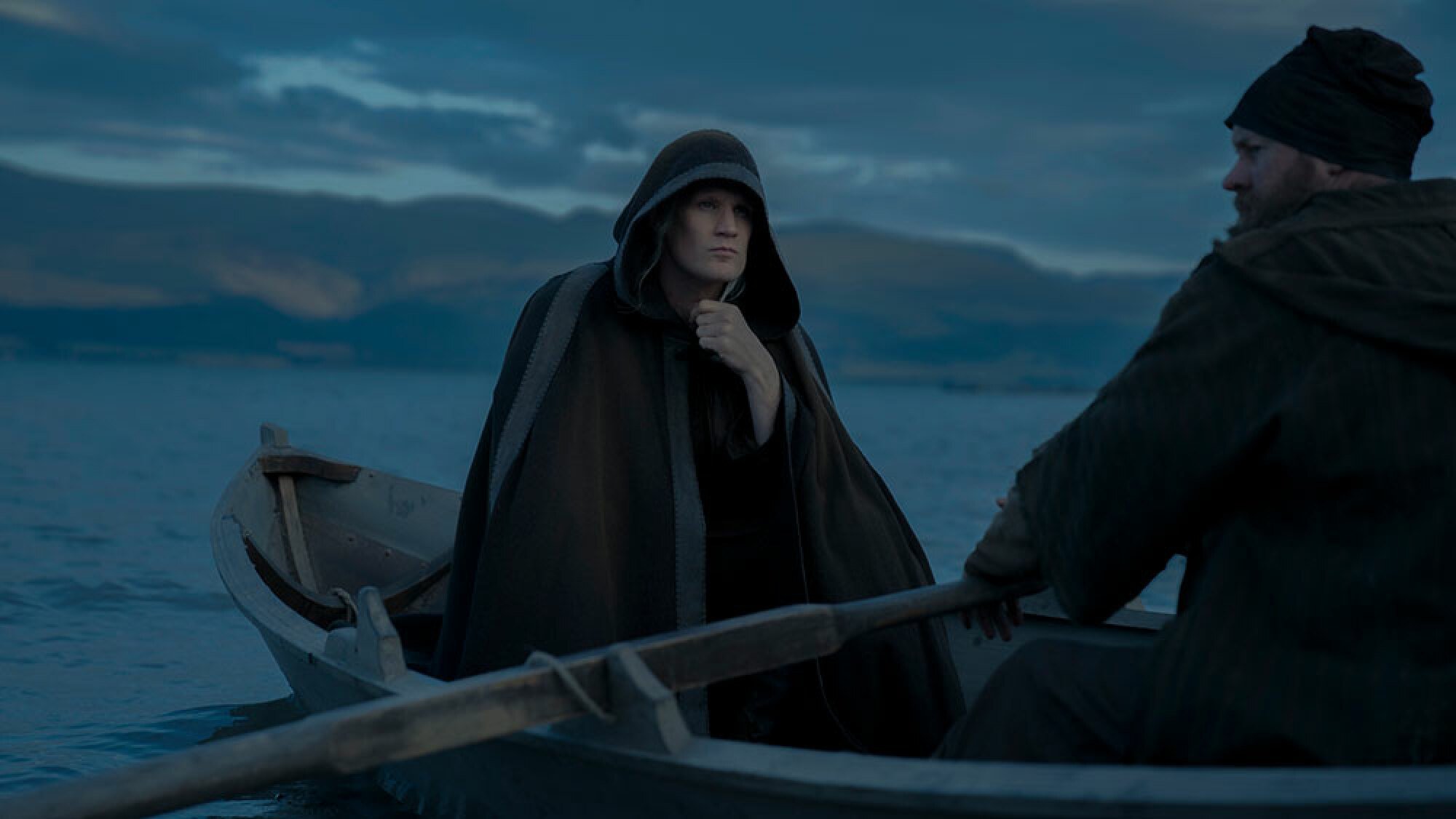 A man in a cloak sits in a small rowing boat while another man rows.