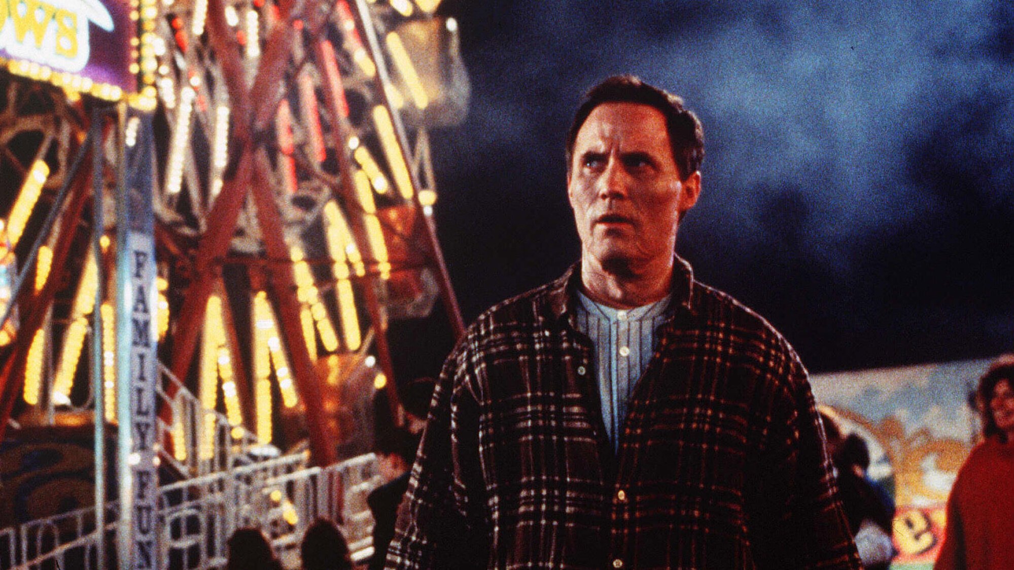 A man stands in a fairground, with a lit-up ferris wheel behind him.