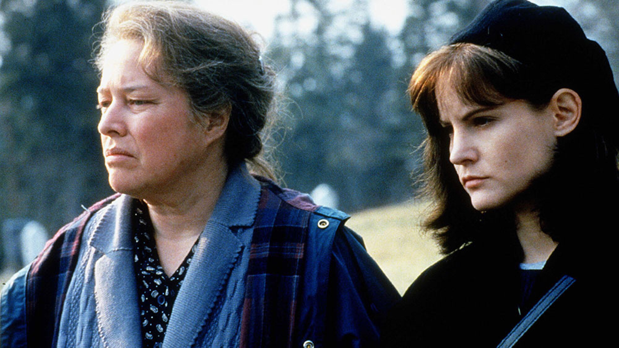 Two women stand looking solemn in a graveyard.